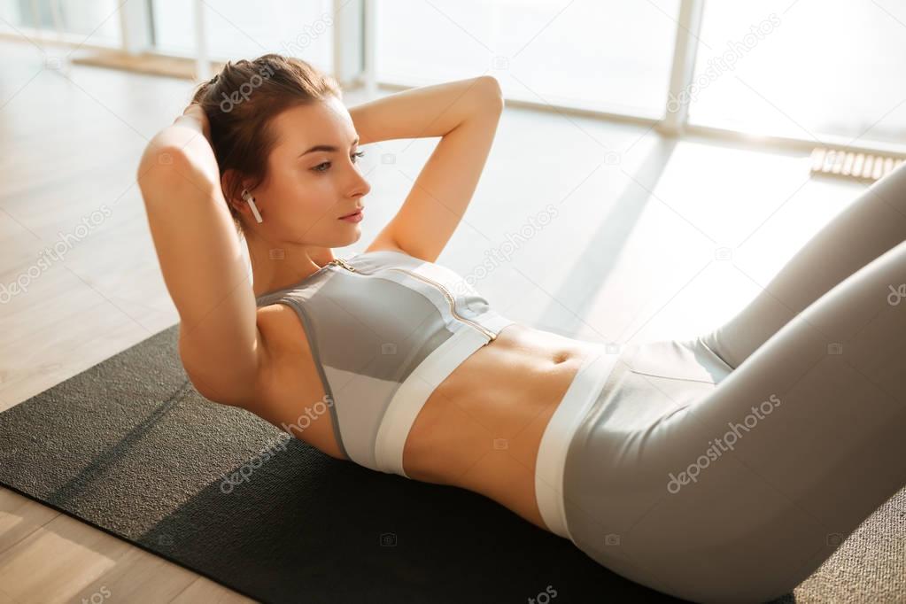 Portrait of thoughtful girl in sporty top and leggings lying on yoga mat and rock press while listening music in earphones at home with big windows on background