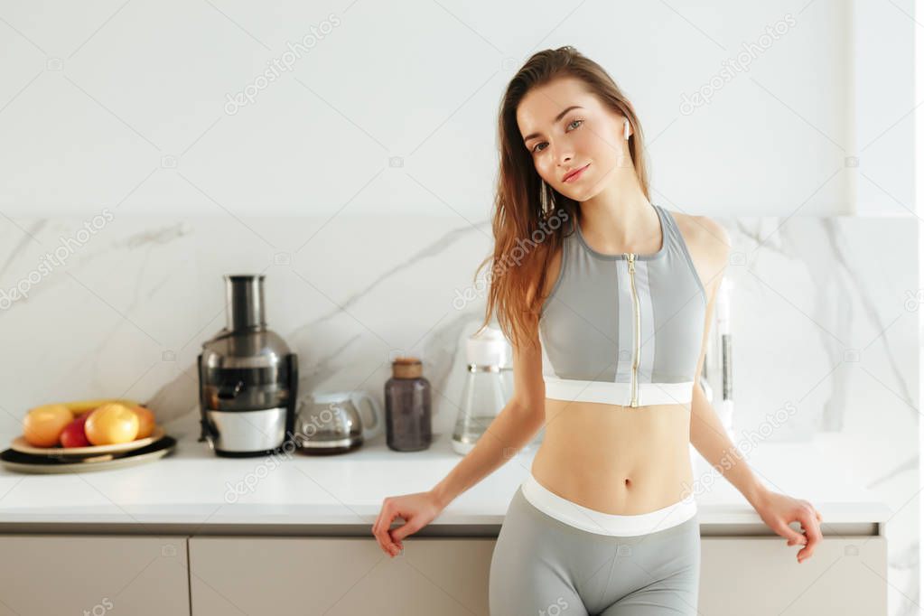 Portrait of beautiful lady in sporty top and leggings standing on kitchen in earphones and dreamily looking in camera isolated