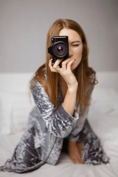 穿着天鹅绒长袍的年轻酷女人的肖像坐在床上 手持黑色的小相机在手隔离 — 图库照片