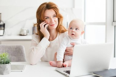 Gülümseyen dizüstü bilgisayar ve cep telefonunu üzerinde şirin küçük bebeği elinde tutarak konuşmak masada oturan iş kadın portresi izole