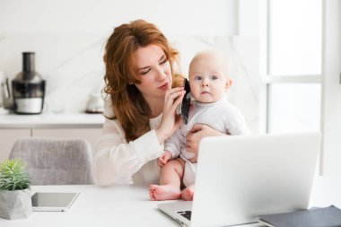 Laptop ile masada oturan ve sevimli küçük bebeği ile oynarken cep telefonu elinde tutan güzel iş kadın portresi izole