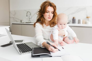 Close up photo of business woman holding her little baby while sitting at the table and making notes in notebook isolated clipart
