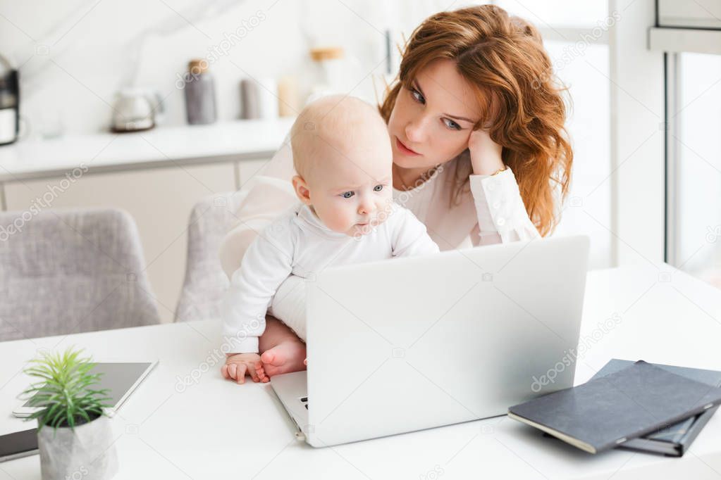 Portrait of tired business woman sitting at the table and working on laptop while holding her little baby near isolated