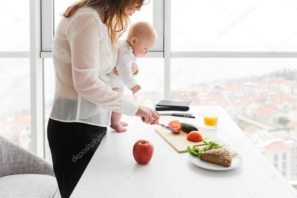 Portrait of young mother standing and holding her little baby while cooking on kitchen with big windows on background