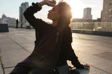 Yarı-yüz Afrikalı kıvırcık kız siiting açık havada cityscape üzerinden günbatımı aydınlatmalı çatıda portresi. Güneşin doğuşunu kentsel arka planda kadın