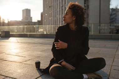 Yarı-yüz Afrikalı kıvırcık kız siiting açık havada almak uzakta kahve içme günbatımı aydınlatmalı çatı üzerinde cityscape portresi. Kıvırcık kadın kentsel arka plan soğuk sabah güneşin doğuşunu üzerinde