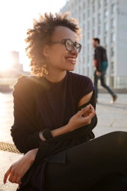 Neşeli Afro-Amerikan kıvırcık kız siiting gün batımı aydınlatmalı sokakta açık havada cityscape üzerinde gülümseyen. Afrikalı kadın kentsel arka plan üzerinde