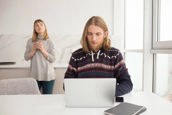 Portrait of young man in sweater sitting at the table and working on laptop while girl on background standing with mug and thoughtfully looking aside