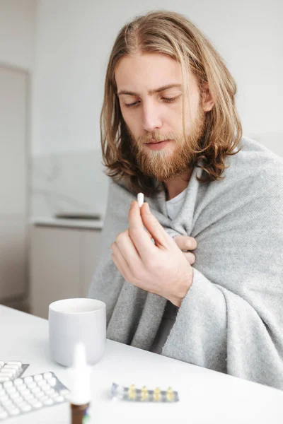 Portrait of young ill man sitting with mug and thoughtfully looking on pill  in hand in kitchen at home isolated