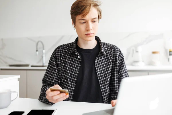 Portrait of young man in earphones sitting with credit card in hand and working on laptop in kitchen at home isolated