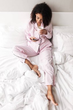 Güzel Bayan pijama Kupası ve cep telefonu elinde evde yatakta oturmuş siyah kıvırcık saçlı izole