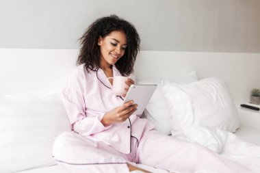 Kupası ve ellerde evde tablet ile yatakta oturmuş pijama siyah kıvırcık saçlı gülümseyen kadın portresi izole