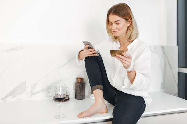 Portrait of young happy lady sitting on kitchen counter with cellphone and credit card in hands and glass of red wine near at home isolated