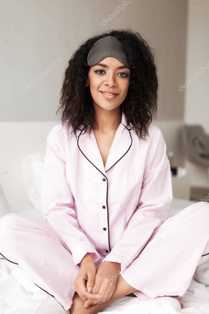 Portrait of beautiful woman with dark curly hair sitting on bed with eye mask on head and happily looking in camera isolated