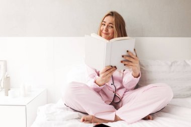 Genç gülümseyen kadın yatakta oturuyor ve sevinçle seyir evde kitap izole portresi