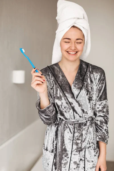 Portrait of cheerful lady in velvet robe standing with towel on head and toothbrush in hand at home isolated