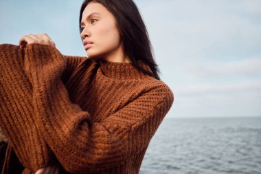 Güzel Asyalı esmer kız, rahat örgü süveter giyer. Düşünceli bir şekilde sahilde poz verir.