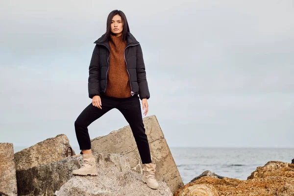 Attraktiv afslappet asiatisk pige i dunjakke kigger i kameraet poserende på store sten ved havet - Stock-foto