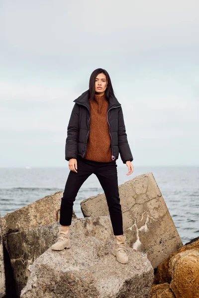 Krásná ležérní asijské dívka v bundě zamyšleně dívá do kamery na velkých kamenech u moře — Stock fotografie