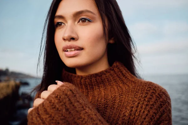 Portrait of pretty Asian brunette girl in knitted sweater dreamily looking away on seaside — Stok Foto