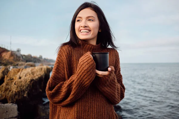 Ganske munter asiatisk brunette jente i strikket genser med krus i hendene ser gladelig bort på havet – stockfoto