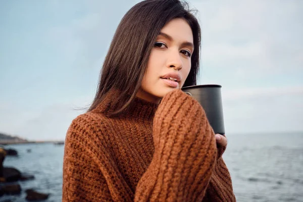 Beautiful Asian brunette girl in knitted sweater with mug in hands intently looking in camera on seaside — Stock Photo, Image