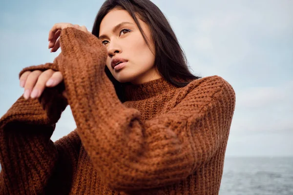 Красивая азиатская брюнетка в уютном трикотажном свитере мечтательно смотрит вдаль, позируя на берегу моря — стоковое фото