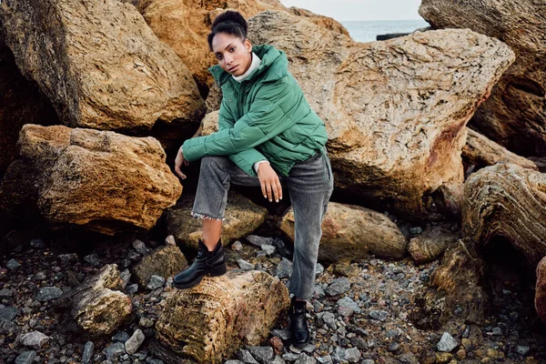 Привлекательная случайная афроамериканка в стильной куртке уверенно смотрящая в камеру на камнях у моря — стоковое фото