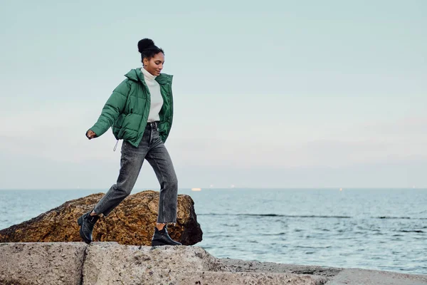 Красивая случайная афроамериканка в стильной куртке мечтательно прогуливаясь по камням у моря — стоковое фото