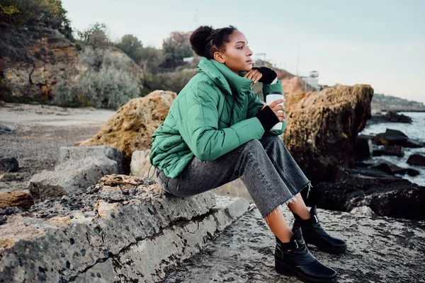 Sidovy över vackra afroamerikanska flicka i dunjacka med kopp för att gå drömskt sitter på stenar vid havet — Stockfoto