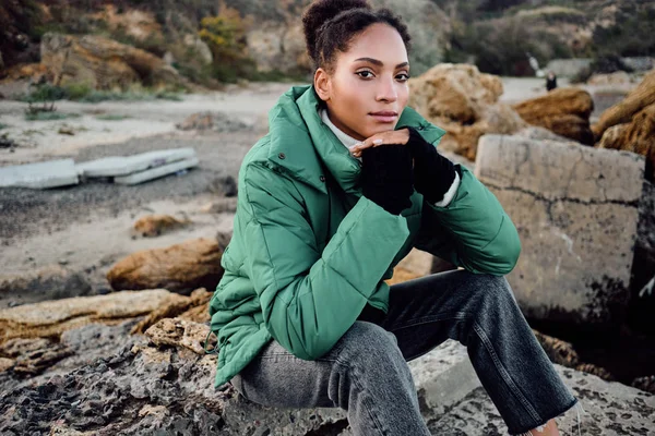 Vacker snygg afroamerikansk flicka i dunjacka tryggt tittar i kameran sitter på stenar utomhus — Stockfoto