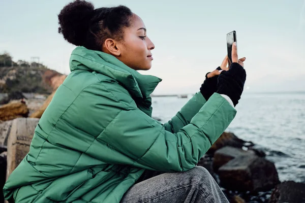 海によって携帯電話で写真を撮るダウンジャケットの笑顔カジュアルなアフリカ系アメリカ人の女の子の側面図 — ストック写真