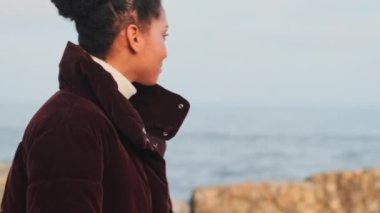 Kendine güvenen Afro-Amerikan kızı yakından çek, ceketli, mutlu bir şekilde deniz kenarında yürüyor.