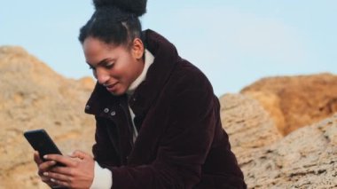 Güzel gülümseyen, sıradan bir Afro-Amerikan kızı ceketli, cep telefonuyla deniz kenarında taşlara mesaj atıyor.