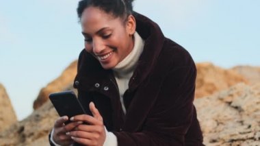 Güzel, Afro-Amerikalı, ceketli, deniz kenarında taşların üzerinde cep telefonuyla gülen bir kız.
