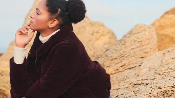 海によって石の上に思慮深くポーズでダウンジャケットの魅力的なアフリカ系アメリカ人の女の子の撮影 — ストック動画