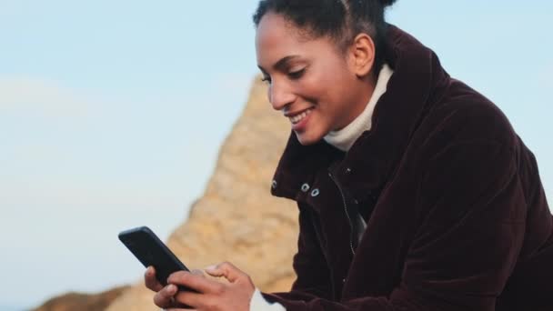 かなりカジュアルなアフリカ系アメリカ人の女の子でダウンジャケット喜んでテキストの携帯電話で石の上に海 — ストック動画