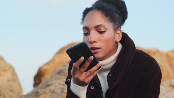 穿着羽绒服的迷人的非洲裔美国女孩在海滨手机上的情感录音留言 — 图库视频影像