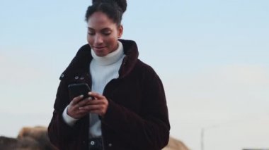 Açık havada cep telefonuyla gezen ceketli Afro-Amerikan bir kızın orta boy fotoğrafı.