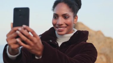 Güzel, Afro-Amerikan bir kız cep telefonuyla fotoğraf çekiyor. V-işareti ve açık hava hareketi gösteriyor.