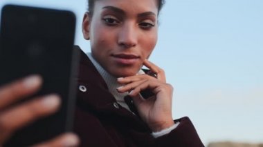 Çekici özgüvenli Afrikalı Amerikalı kız cep telefonuyla selfie çekiyor ve dışarıda gülümsüyor.