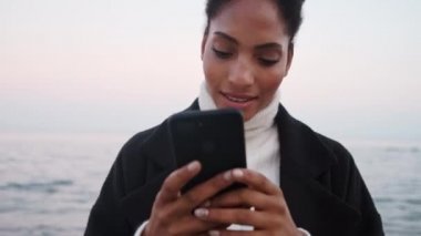 Güzel gülümseyen Afrikalı Amerikalı kız deniz kenarında dinlenen cep telefonunu neşeyle kullanıyor. 