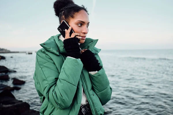 魅力的なアフリカ系アメリカ人の女の子の肖像-ダウンジャケットで携帯電話で海のそばを歩く — ストック写真
