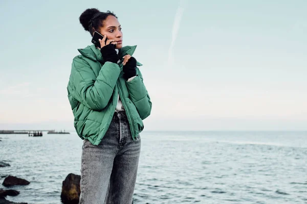 Vacker avslappnad afroamerikansk flicka i dunjacka tankeväckande talar på mobiltelefon vid havet — Stockfoto