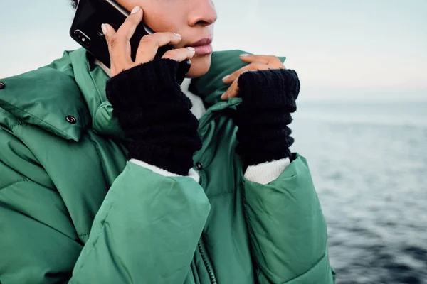 Zamknij się swobodnie Afroamerykanka dziewczyna w dół kurtka mówi przez telefon komórkowy nad morzem — Zdjęcie stockowe