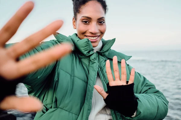 Portret bardzo radosnej Afroamerykanki w kurtce na dół szczęśliwie ciągnącej ręce do kamery nad morzem — Zdjęcie stockowe
