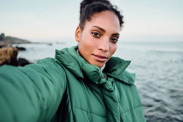 Piękne Afroamerykanka w dół kurtka robi selfie nad morzem — Zdjęcie stockowe