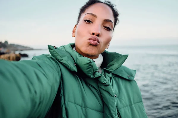 Piękne Afroamerykanka w dół kurtka wysyłanie powietrza pocałunek biorąc selfie nad morzem — Zdjęcie stockowe