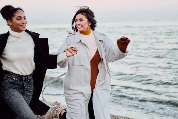 Dua gadis cantik tersenyum gaya dalam mantel parit gembira berjalan oleh laut — Stok Foto