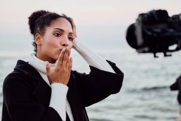 Ganska avslappnad afroamerikansk flicka gladeligen skicka luft kyss på kameran under fotografering vid havet — Stockfoto